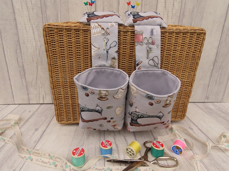 May include: Two grey fabric hanging storage containers with a sewing machine print. The containers are hanging on a wicker basket. There are spools of thread and scissors on a wooden surface.