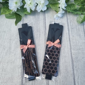 May include: Two black and rose gold bookmark sets with a pink ribbon. The bookmarks have different patterns, one with a geometric pattern and the other with a striped pattern.