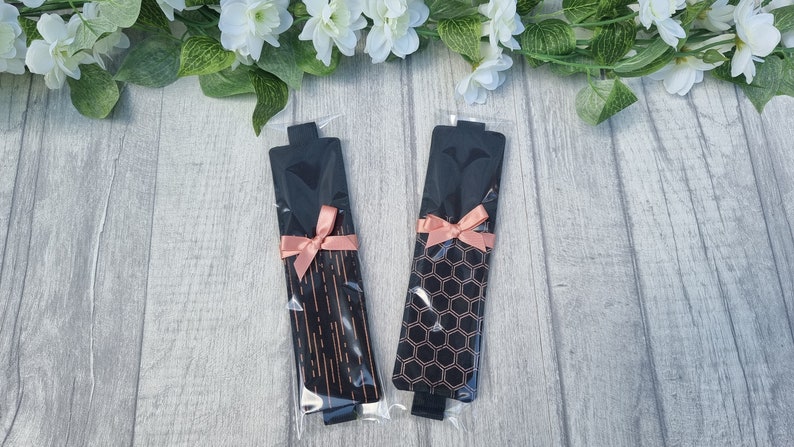 May include: Two black and rose gold bookmark sets with a pink ribbon. The bookmarks have different patterns, one with a geometric pattern and the other with a striped pattern.