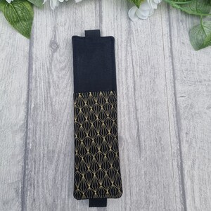 May include: A black fabric bookmark with a gold geometric pattern. The bookmark has a black flap on the top and bottom.
