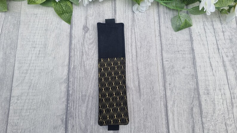 May include: A black fabric bookmark with a gold geometric pattern. The bookmark has a black flap on the top and bottom.