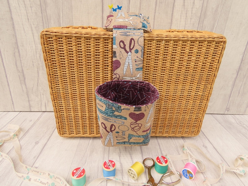 May include: A wicker basket with a fabric hanging organiser. The organiser has a brown and beige fabric with a pattern of sewing tools and a burgundy lining. The organiser is hanging from the top of the basket. There are spools of thread and a pair of scissors on the table in front of the basket.