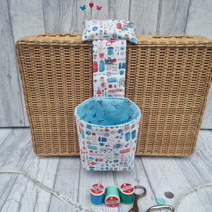 May include: A white wicker basket with a blue and white fabric hanging organiser with a sewing theme. The organiser has a pocket on the top and a hanging bin below. The bin is filled with sewing notions, including scissors, thread, and buttons. There are three spools of thread on the table below the basket.