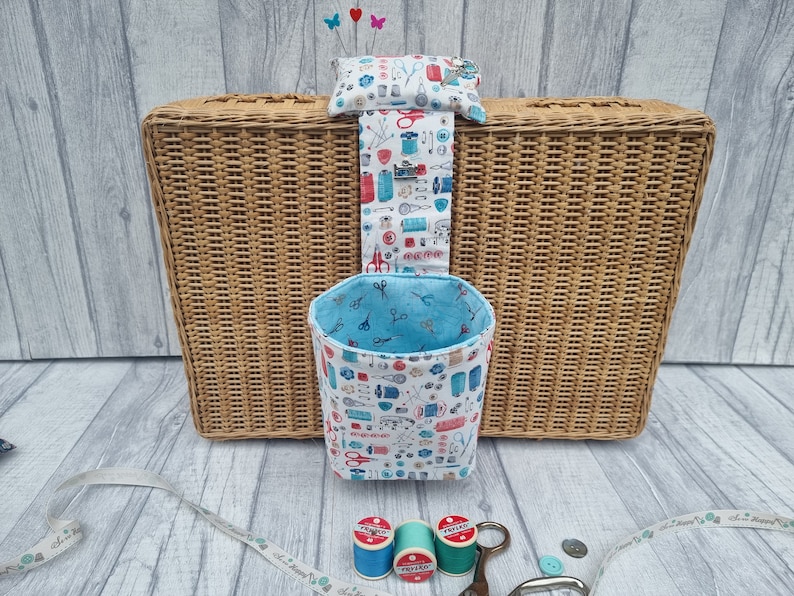 May include: A white wicker basket with a blue and white fabric hanging organiser with a sewing theme. The organiser has a pocket on the top and a hanging bin below. The bin is filled with sewing notions, including scissors, thread, and buttons. There are three spools of thread on the table below the basket.