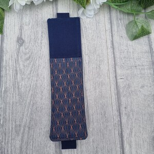 May include: A navy blue fabric phone case with a geometric pattern in copper. The case is closed and lying on a light gray wood surface.