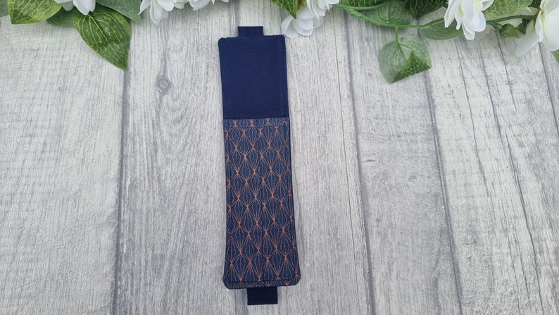 May include: A navy blue fabric phone case with a geometric pattern in copper. The case is closed and lying on a light gray wood surface.