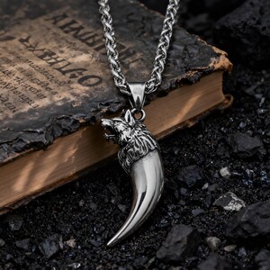 May include: A silver-colored pendant necklace featuring a wolf head and a large claw-shaped design. The necklace has a thick chain and is displayed on a dark, textured surface. The wolf's head is detailed with open mouth and fur.