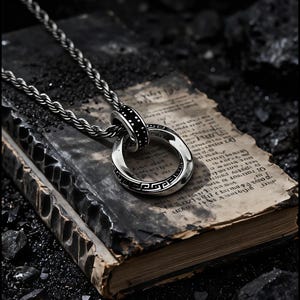 Op de afbeelding: Een zilverkleurige hangerketting met een cirkelvormig ontwerp, een zwarte band en een Grieks sleutelpatroon. De ketting ligt op een open boek met verouderde pagina's, tegen een donkere achtergrond.
