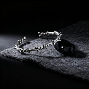 May include: A silver-toned open cuff bracelet featuring a textured design that mimics twisted barbed wire with sharp points. A single, large, faceted black gemstone is attached to one end of the metal band, creating a stark contrast in color.