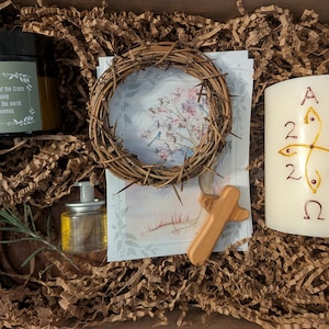 May include: A gift box filled with religious items. Includes a candle with a cross design and the numbers 2026, a wooden cross, a crown of thorns, a jar with a label, and a small bottle of oil. The box is lined with brown paper.