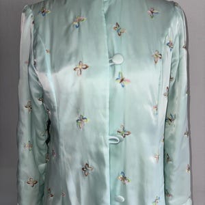 Può includere: Una vestaglia in raso azzurro con collo alla coreana e maniche lunghe. La vestaglia è decorata con farfalle ricamate in vari colori. Presenta una chiusura frontale con bottoni e un bordo in raso abbinato.