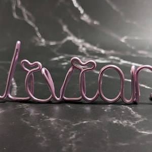 May include: A pink wire name sign spelling the name "Vivian".