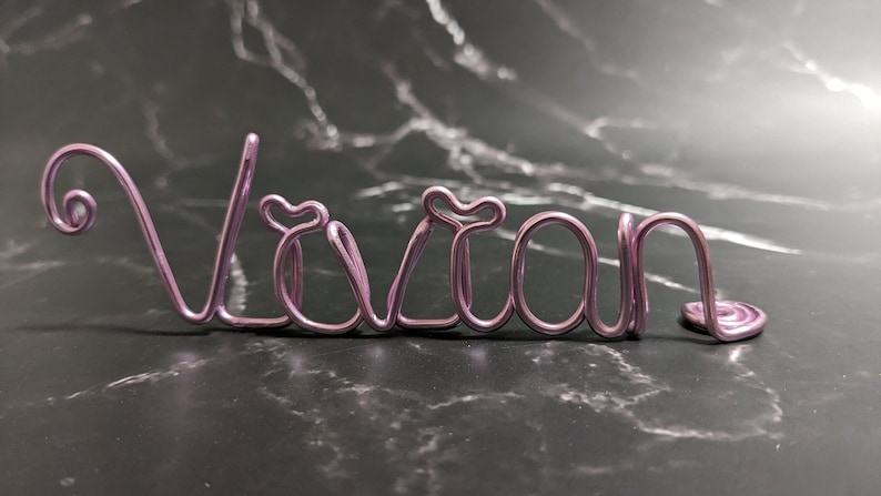 May include: A pink wire name sign spelling the name "Vivian".