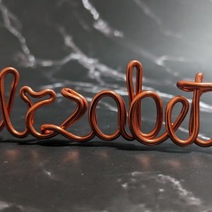 May include: Copper wire sculpture of the name "Elizabeth" in a cursive font. The sculpture is on a dark gray marble surface.