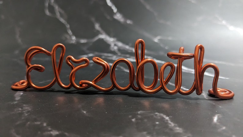 May include: Copper wire sculpture of the name "Elizabeth" in a cursive font. The sculpture is on a dark gray marble surface.