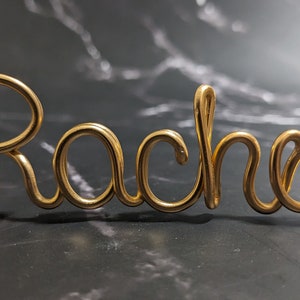 May include: Gold wire sculpture of the name "Rachel" in a cursive font.