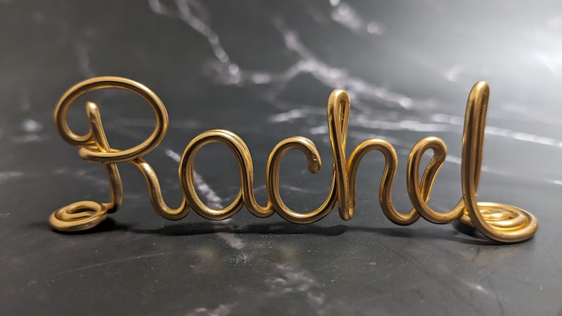 May include: Gold wire sculpture of the name "Rachel" in a cursive font.