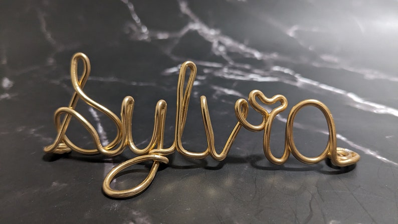 May include: Gold wire sculpture of the name "Sylvia" on a black marble background.