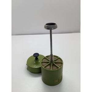 以下が含まれることがあります： 緑色と金属製のヴィンテージキッチンツール。おそらくフードプレスまたはマッシャー。格子模様の円筒形のベース、バネ付きの中央ロッド、円形のトップが特徴です。蓋も緑色で、黒いノブが付いています。
