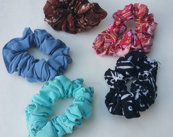 May include: Five different hair scrunchies in various colors and patterns. The scrunchies are made of fabric and have a gathered, ruffled look. The colors include blue, turquoise, pink, brown, and black.