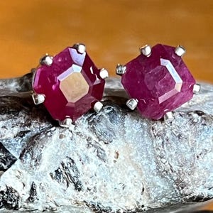 Puede incluir: Un par de pendientes de botón de rubí. Cada pendiente presenta una gema facetada de color rojo intenso engastada en metal plateado. Los pendientes se exhiben sobre una superficie de piedra gris y blanca.