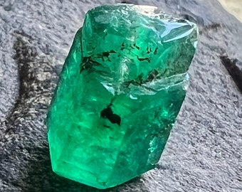 May include: A large, faceted emerald gemstone with a deep green color. The gemstone is cut into a rectangular shape with sharp edges and a smooth, polished surface.