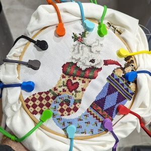 May include: A close-up of a cross-stitch project depicting a Christmas stocking with a snowman and a reindeer. The project is mounted in a wooden embroidery hoop and secured with colorful clips.