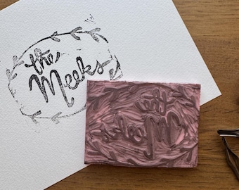 May include: A custom stamp with the words "the Meeks" in a script font, surrounded by a floral wreath design. The stamp is next to a paper with the stamped image. The stamp is pink and the stamped image is black.