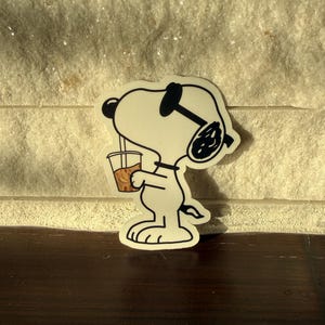 May include: A die-cut sticker featuring a white cartoon dog with black ears and a black collar, wearing dark sunglasses. The character is holding a clear plastic cup filled with a light brown iced beverage and a straw, rendered in a simple line art style.