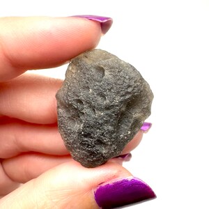 May include: A dark gray, irregularly shaped stone with a rough, textured surface. The stone is held between fingers with purple nail polish, against a white background. The stone appears to be a natural mineral or crystal.