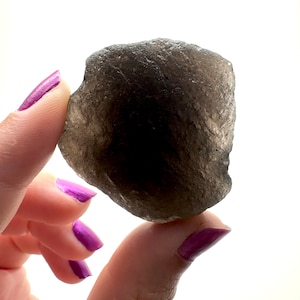 May include: A dark gray smoky quartz crystal with a rough, irregular shape. The crystal is being held in a hand with purple nail polish.