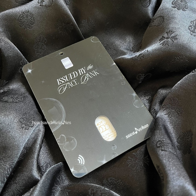 May include: A rectangular black card featuring the text ISSUED BY the FACE BANK, beadsandmelodies, and sawaDeehao. The card includes a silver chip, a contactless payment symbol, and a central cutout revealing a gold-toned circular emblem.