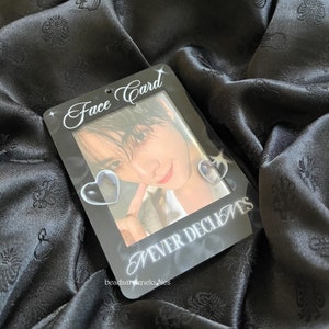 May include: A rectangular black acrylic photo holder featuring the white text Face Card at the top and NEVER DECLINES at the bottom. The frame includes two small heart-shaped cutouts on the sides and displays a portrait of a person.
