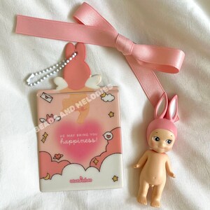 Op de afbeelding: Een roze en witte kaart met een konijnenontwerp en de tekst "He may bring you happiness!" De kaart is bevestigd aan een zilveren ketting en heeft een roze lint eromheen. Een klein roze konijnenbeeldje is ook op de afbeelding te zien.