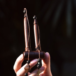May include: A dark brown wooden musical instrument with two vertical tubes connected to a horizontal base. The instrument has a string and decorative beads. The background is dark.