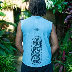 May include: A sleeveless denim shirt with a black and white graphic print on the back. The graphic features a snake, a bird, and a human figure. The shirt is light blue and has a collar.