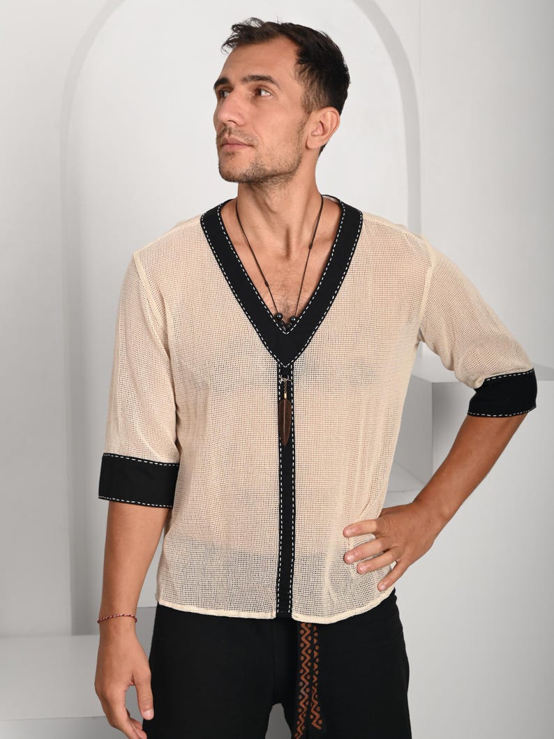 May include: A beige mesh shirt with black trim around the neckline and sleeves. The shirt has a V-neck and a black cord with a wooden pendant hanging from the neckline.