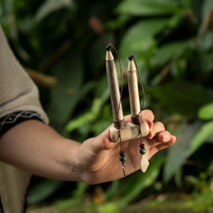 May include: A handcrafted wooden musical instrument, held in a hand against a blurred green background. The instrument features two vertical wooden pieces with dark string and small black beads, connected to a horizontal base. Decorative green beads and a diamond-shaped pendant hang from the base.
