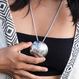 Può includere: Una collana con pendente in metallo argentato martellato. Il pendente è un grande cerchio strutturato sospeso da una delicata catena. La collana è indossata sopra un top nero e uno scialle fantasia.
