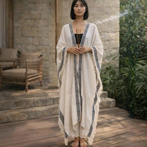 May include: An off-white, floor-length open-front kimono featuring a dark blue geometric pattern along the front edges and hem. The garment has wide sleeves and long fringe detailing along the bottom edge, worn over a black top and light pants.
