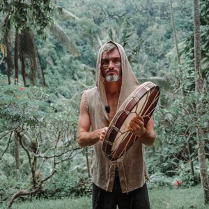 May include: A person wearing a hooded, sleeveless shirt holds a large, round drum. The drum has a brown wooden frame and a light-colored, patterned surface. The background features lush green foliage.