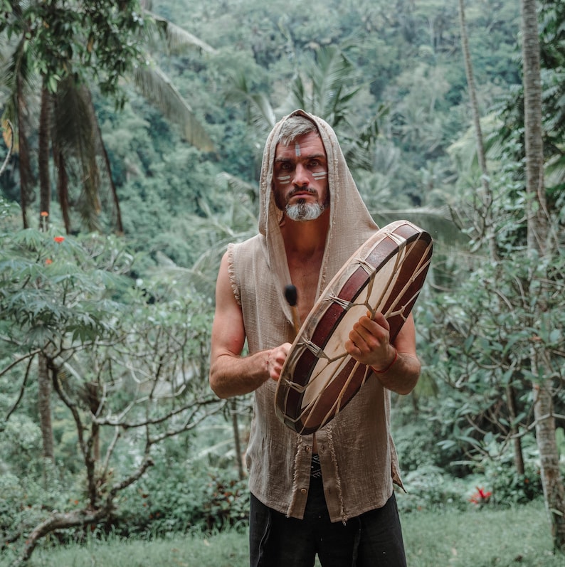 May include: A person wearing a hooded, sleeveless shirt holds a large, round drum. The drum has a brown wooden frame and a light-colored, patterned surface. The background features lush green foliage.