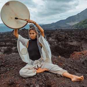 May include: A woman wearing a white robe and black pants sits cross-legged on a rocky surface, holding a large wooden drum overhead with a stick.