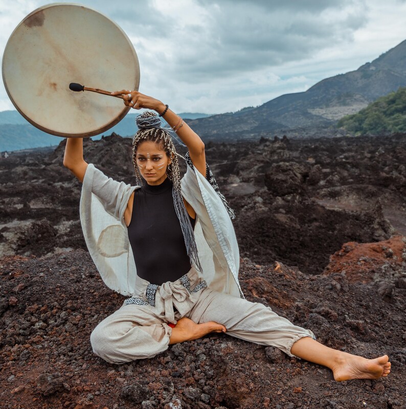 May include: A woman wearing a white robe and black pants sits cross-legged on a rocky surface, holding a large wooden drum overhead with a stick.