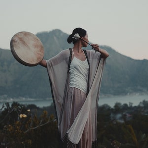 Op de afbeelding: Een vrouw in een wit topje en een beige kimono met zwarte bies houdt een ronde houten trommel voor zich. Ze staat op een stenen platform met een bergketen op de achtergrond.