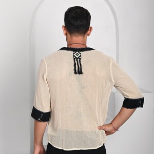 May include: A beige mesh shirt with black trim and a black and white geometric design on the back. The shirt has a tassel hanging from the design.