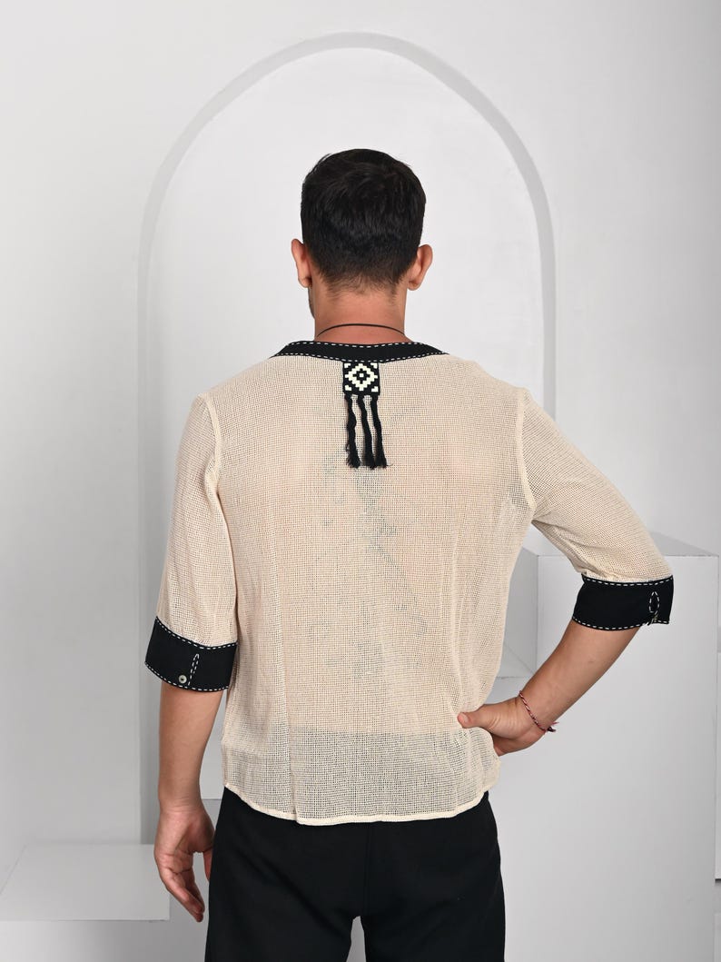 May include: A beige mesh shirt with black trim and a black and white geometric design on the back. The shirt has a tassel hanging from the design.