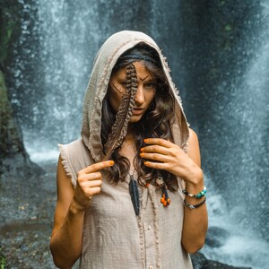 May include: A person wearing a beige hooded sleeveless top with a feather accessory. The person has orange nail polish and a black headband. A waterfall is in the background. The person is wearing a necklace with a feather pendant and bracelets.