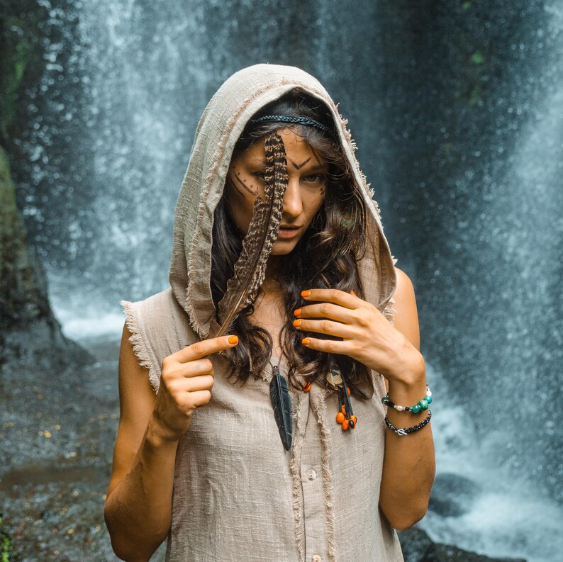 May include: A person wearing a beige hooded sleeveless top with a feather accessory. The person has orange nail polish and a black headband. A waterfall is in the background. The person is wearing a necklace with a feather pendant and bracelets.