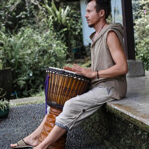 May include: A person playing a djembe drum outdoors. The drum is brown with vertical carved lines and a dark drumhead. The person is wearing a sleeveless tan shirt, light gray pants, and sandals. The background includes lush green foliage and a building.